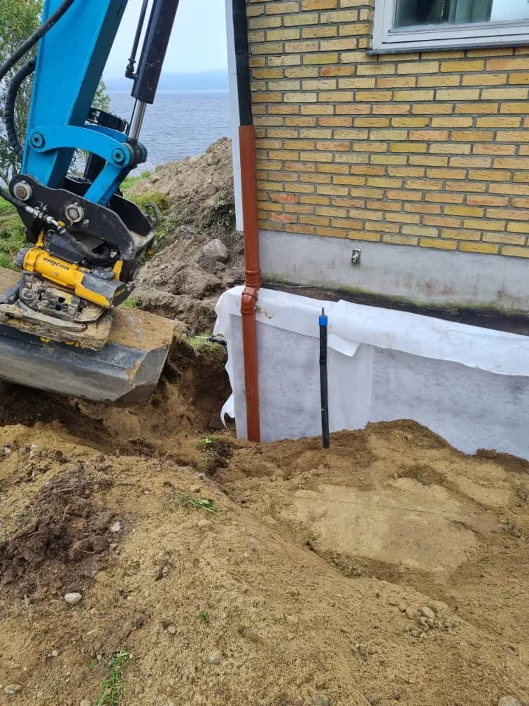 Blå grävmaskin med engcon tiltrotator gräver längs grunden på ett gult tegelhus, med fiberduk mot väggen och havet i bakgrunden