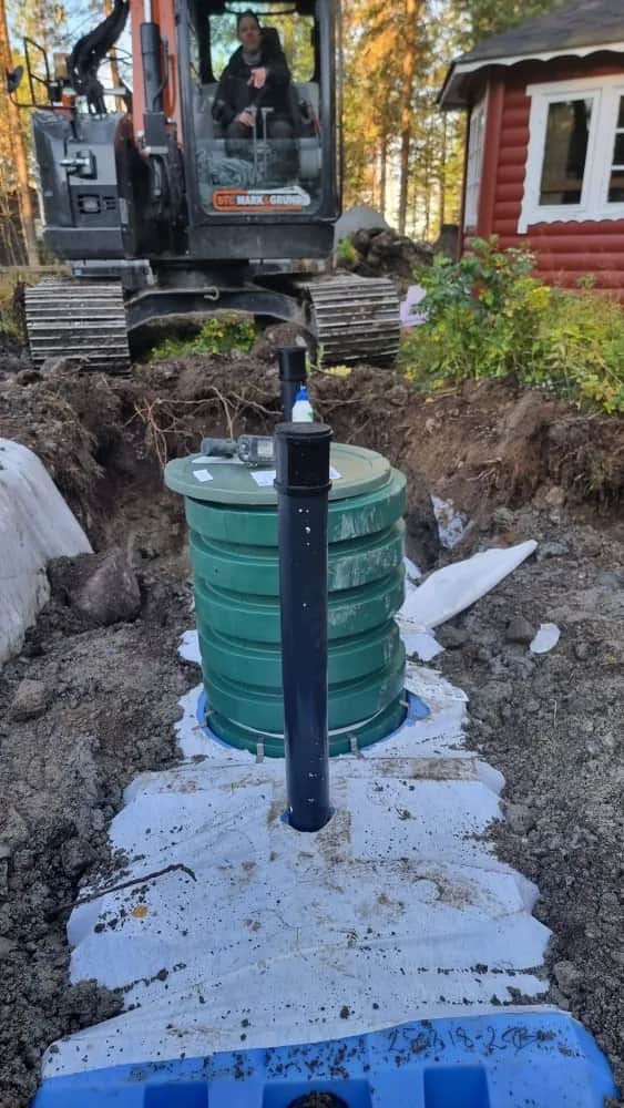 Grön pumpbrunn med svart stigarrör står på fiberduk i schakten framför en Hitachi-grävmaskin och ett rött timmerhus