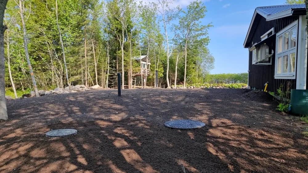 Svart hus med vita knutar vid vattnet, med nyjämnad grusyta och två brunnslock framför