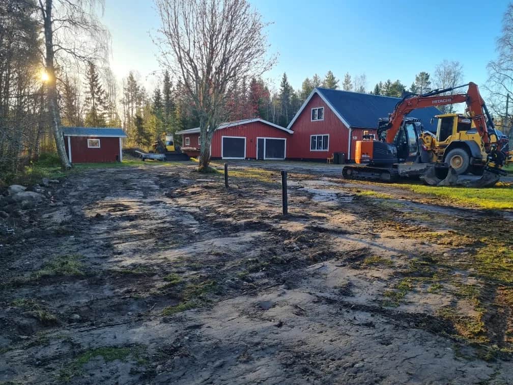 Gårdsplan med röda hus där en Hitachi-grävmaskin och en gul hjullastare står i lågt höstljus