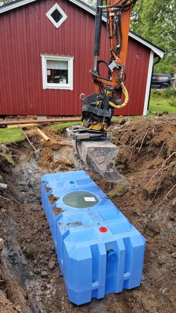 Blå slamavskiljare i schakten intill ett rött hus, med grävmaskinens tiltrotator och skopa ovanför