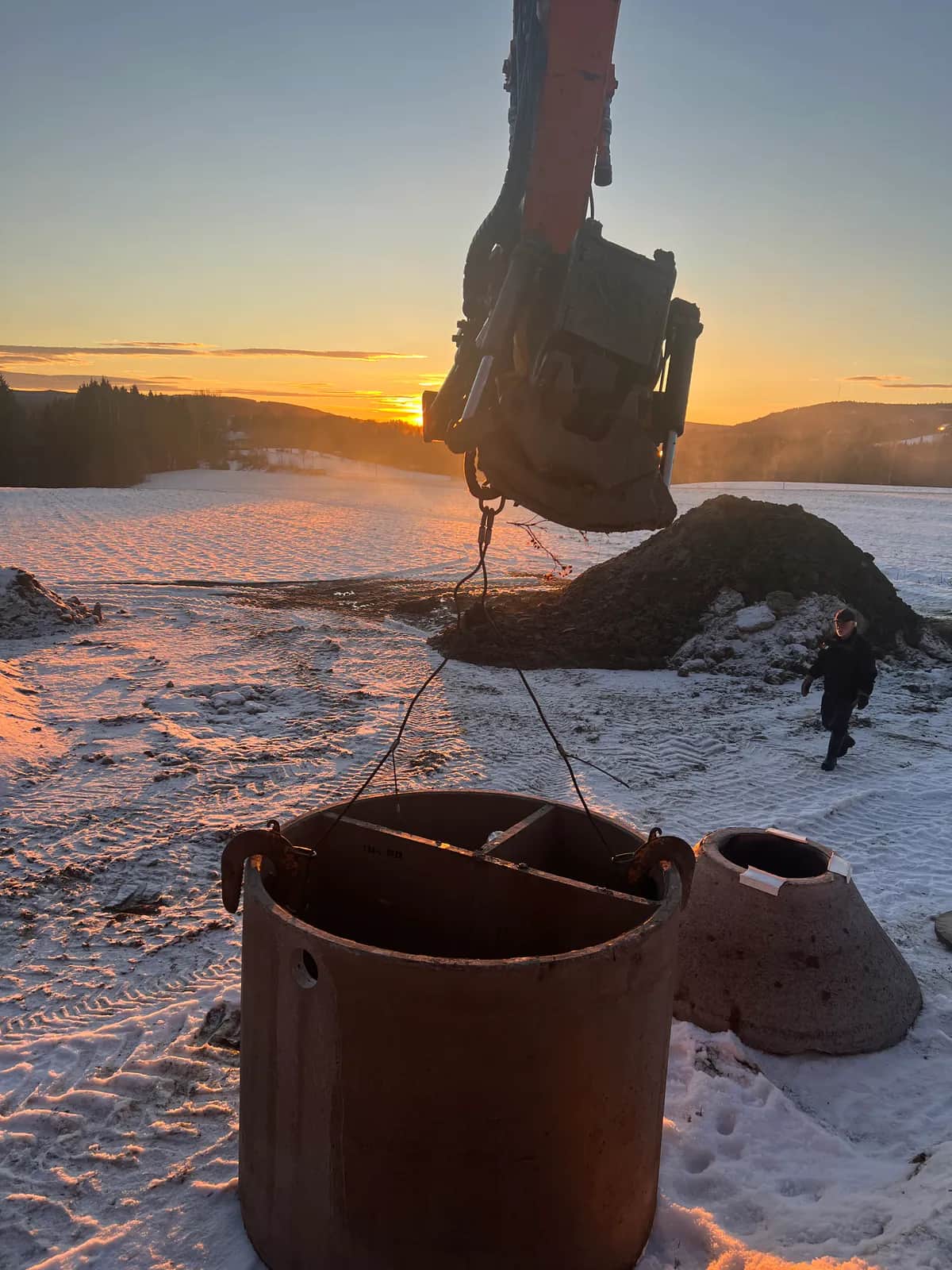 Grävmaskinen lyfter bottendelen till en FQB-betongbrunn i kedjor över snötäckt mark i soluppgången, med den koniska toppen bredvid