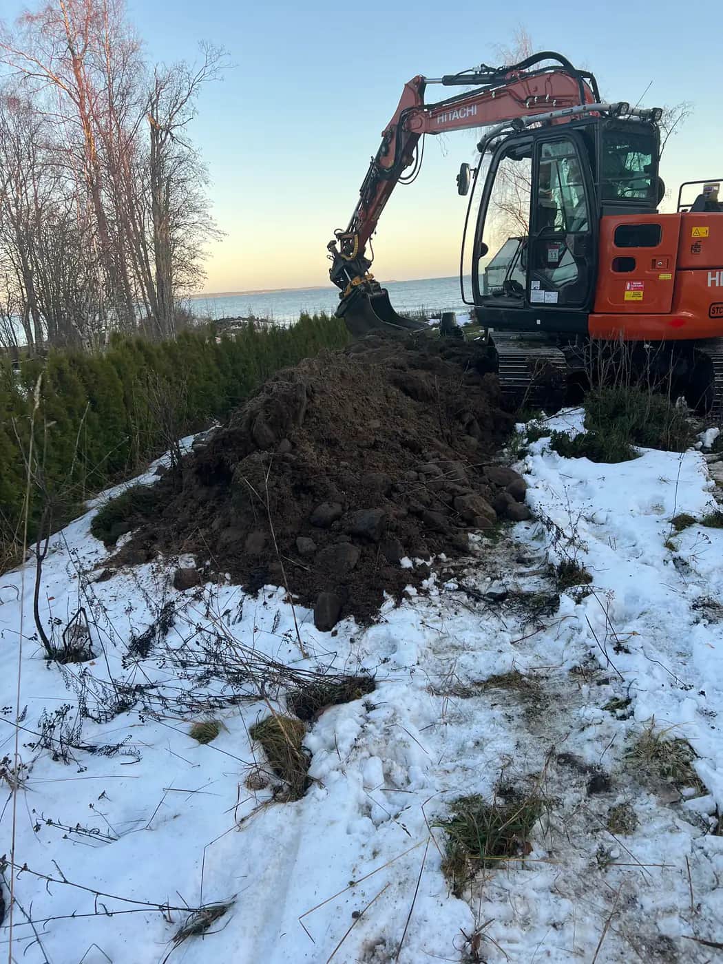 Orange Hitachi bandgrävare gräver en schakt intill en häck med havet och snötäckt mark i bakgrunden en klar vinterdag