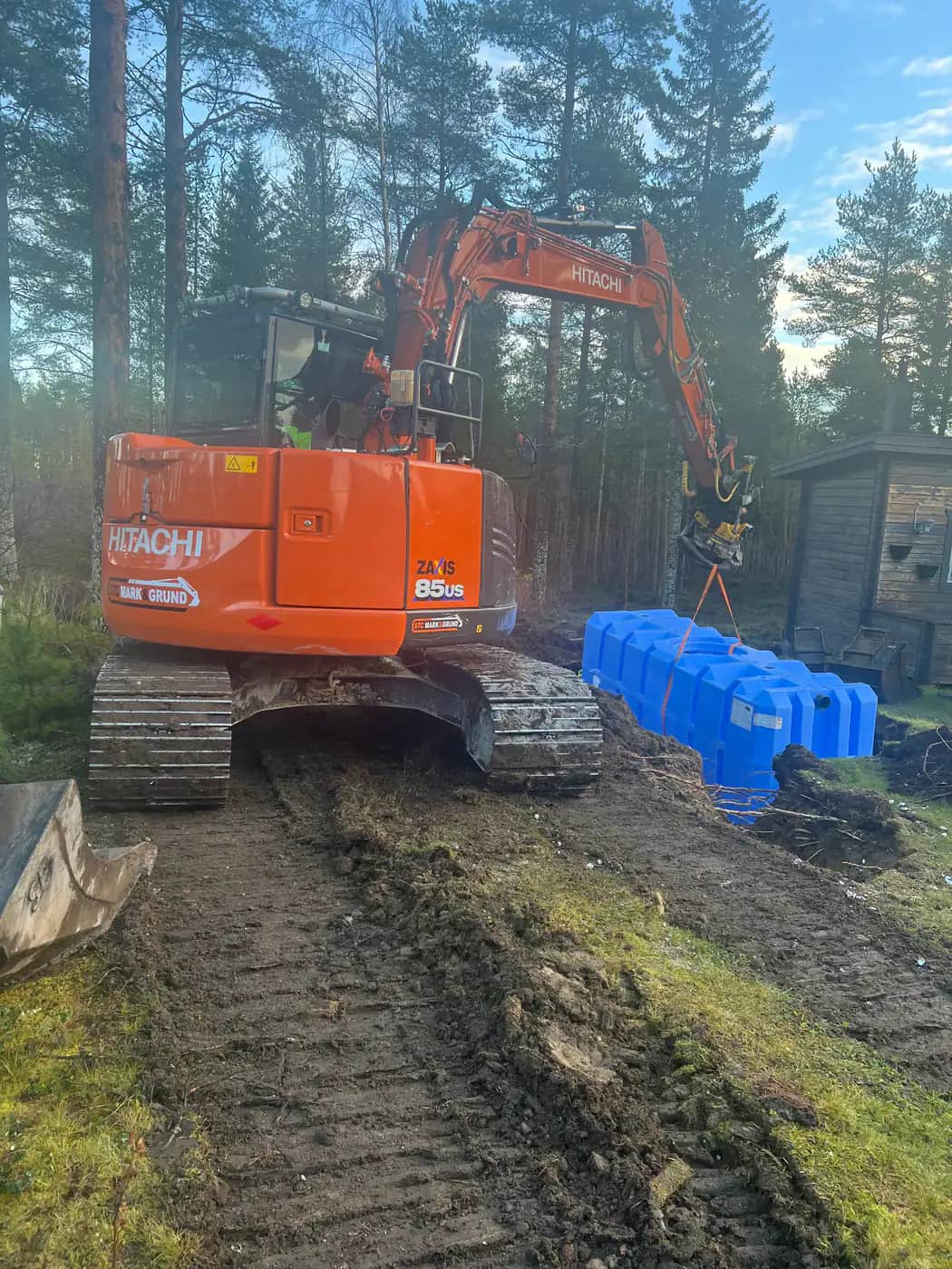 Orange Hitachi ZX85US bandgrävare lyfter en blå slamavskiljare i stroppar på en skogstomt med tallar och ett uthus i bakgrunden