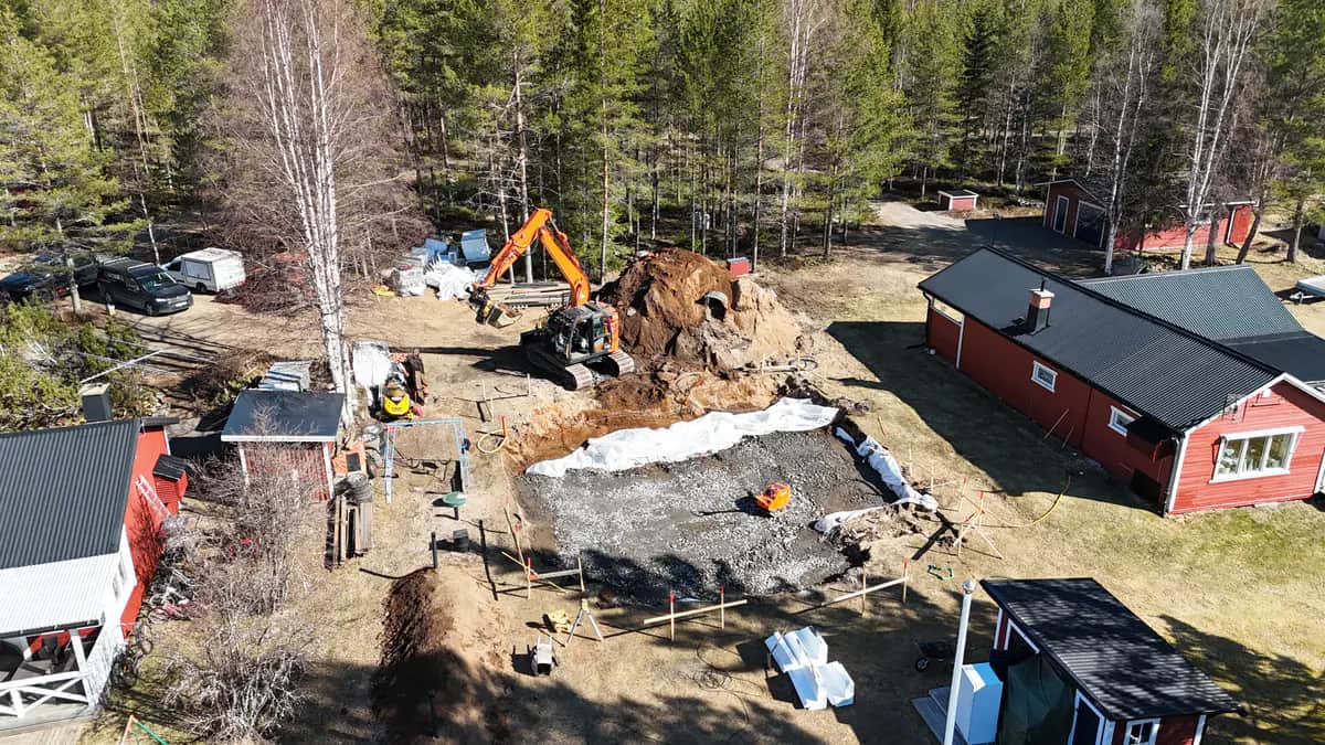 Drönarbild över tomt i skogskant där en grävmaskin står vid en jordhög intill en packad grusbädd för platta på mark, med röda hus runt om