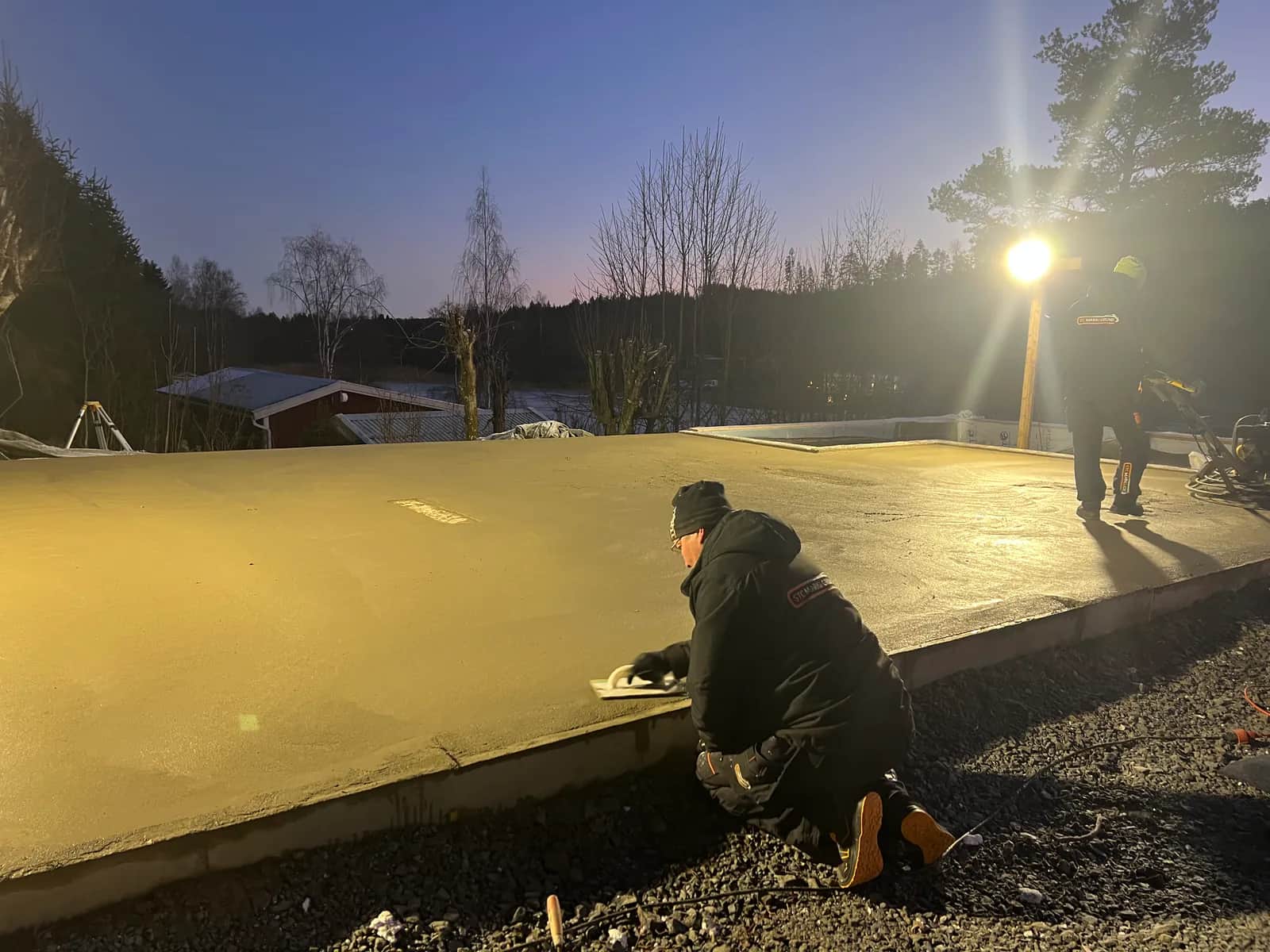 Nygjuten platta på mark i skymningen med en person som stryker kanten och arbetslaget vid strålkastaren i bakgrunden